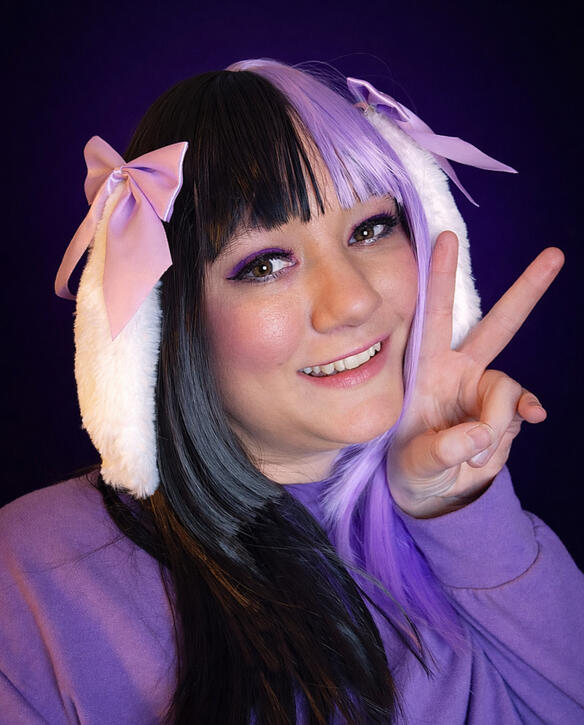 Portrait of digital creator Cinnabunnyr0ll with split-dye purple and black hair. She is wearing a purple sweater, lavender ribbons, and white fluffy bunny ears while smiling and making a peace sign against a deep purple background.